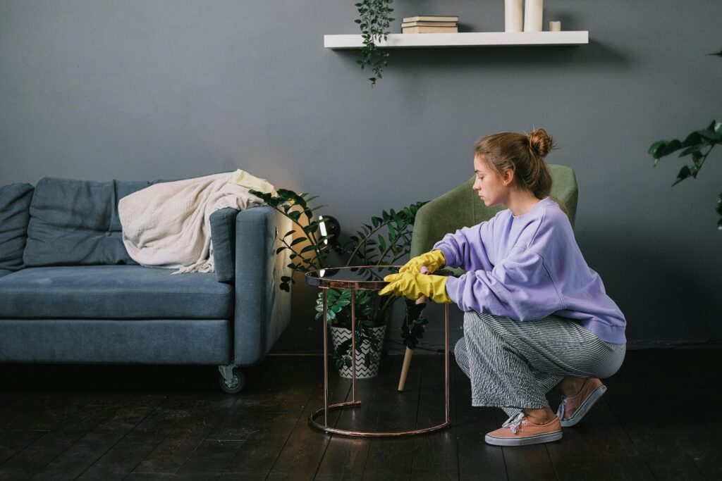 Side view of young female in latex gloves squatting down and wiping table in stylish living room in daytime